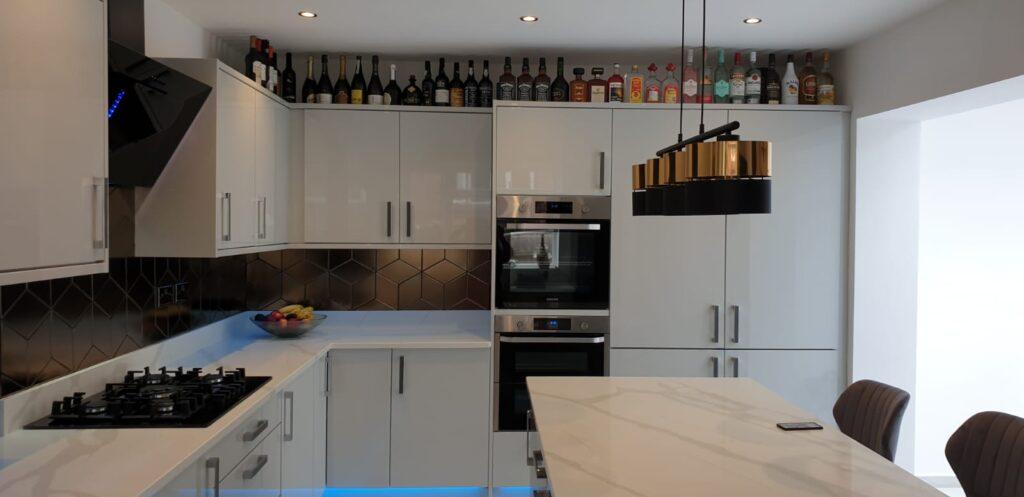 Kitchen with island in foreground and light coloured cupboards in background with bottles above